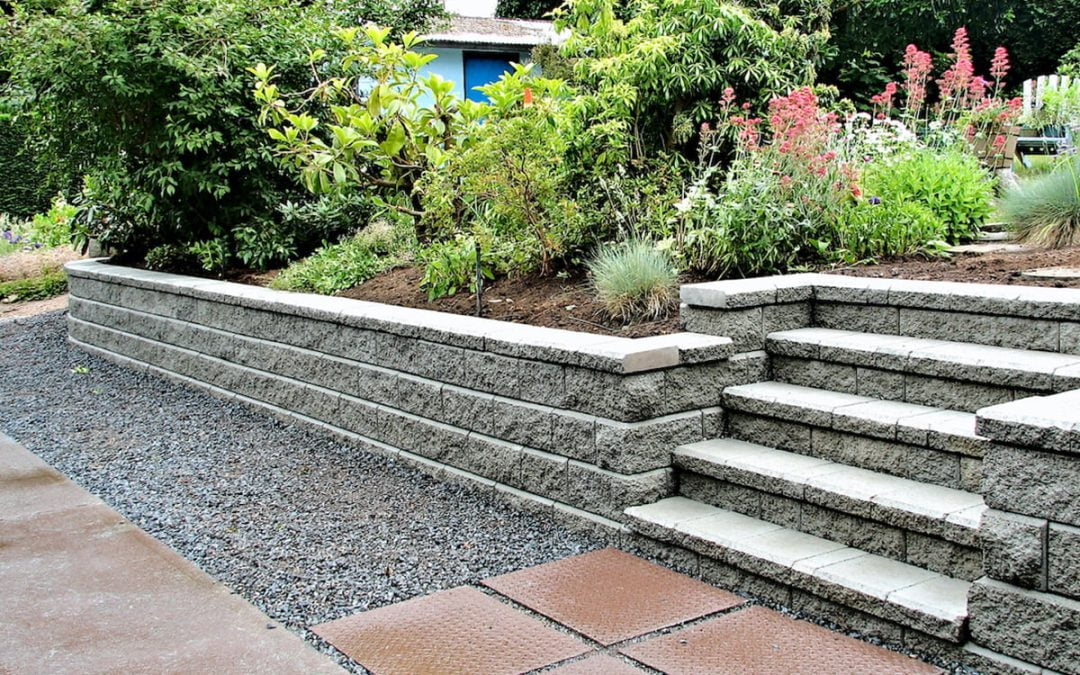 Stone retaining wall with steps, surrounded by lush greenery and blooming plants. Gravel and paved path on the left.